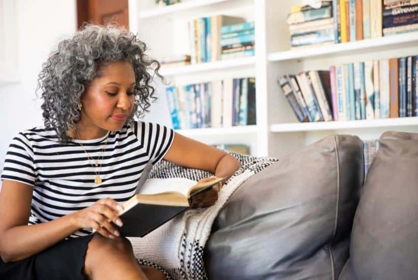 a woman wearing a striped shirt sitting on a silver sofa reading a book with bookshelves in the background featured image for Hope in the Midst of Loss Best Books About Heaven and Loss