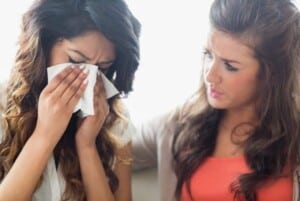 a woman wearing a white shirt who's crying and wiping her tears away with a tissue while another woman wearing a red shirt has her hard on her shoulder trying to comfort her. featured image for How to Comfort Someone Who is Grieving in 5 Practical Ways