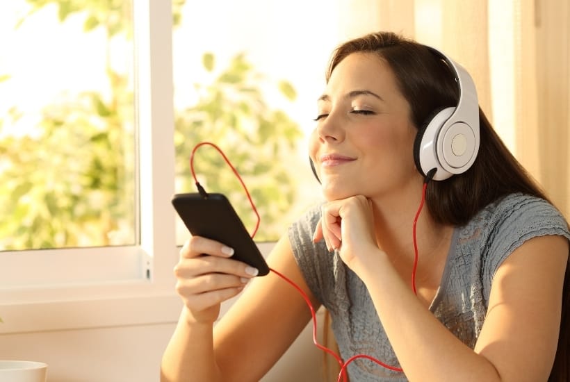 a brown haired woman wearing a gray t-shirt with white headphones on listening to music featured image for The Perfect Christian Playlist For Summer 2021