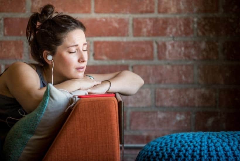 a woman with with headphones on with her arms crossed laying on a burnt orange couch with a brick background featured image for The Ultimate List of Grief Podcasts for 2022