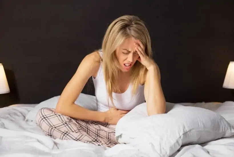 a girl sitting on a bed wearing plaid pj's and white tank top holding her stomach with one hand and her forehead with the other featured image for Why Does God Allow Pain and Suffering?